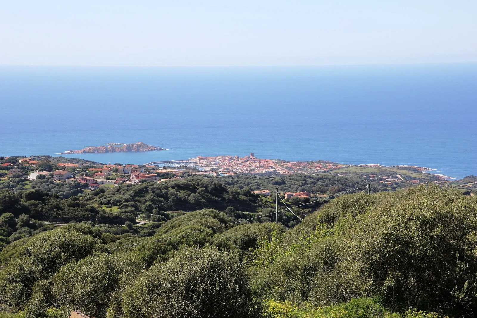 Veduta dall'alto di Isola Rossa con il porto, la torre costiera e l'isolotto di roccia rossa nel mare, tra colline di macchia mediterranea